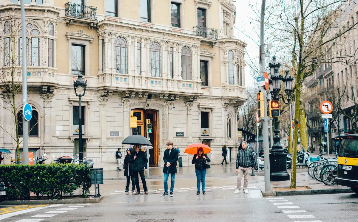 People with umbrellas crossing a rainy street in front of a historic building in Barcelona.