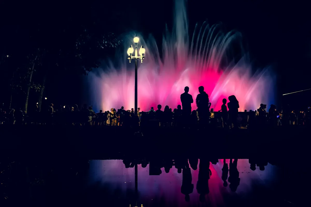 Silhouetted crowd at the vibrant Magic Fountain in Barcelona, with colorful water displays at night.